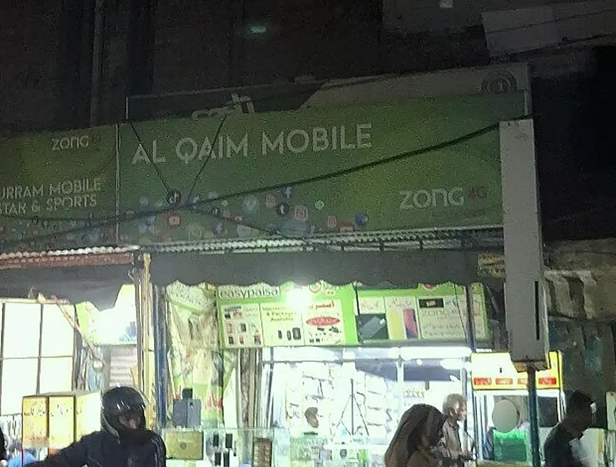 The Q Block shop row at night: the green Al Qaim Mobile board over lit counters, beside Khurram Mobile